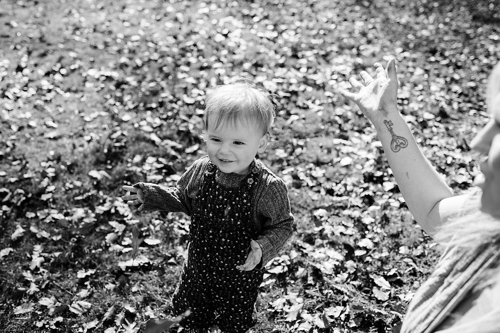 Toddler in patterned overalls standing on leafy ground, smiling. An adult's arm with a tattoo is raised nearby. Black and white image. Family documentary photography Durham and North East. Angela Fenwick Photography
