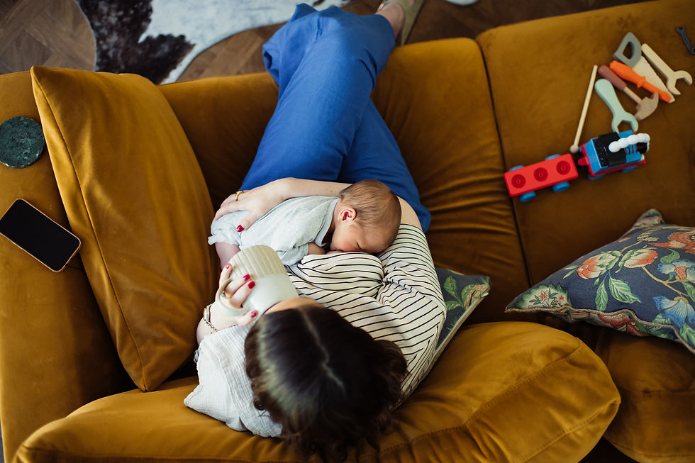 Woman cradling a baby on a mustard sofa, holding a mug. Blue pants, striped shirt. Toys and floral pillows nearby. Natural newborn shoots at home | Angela Fenwick Photography