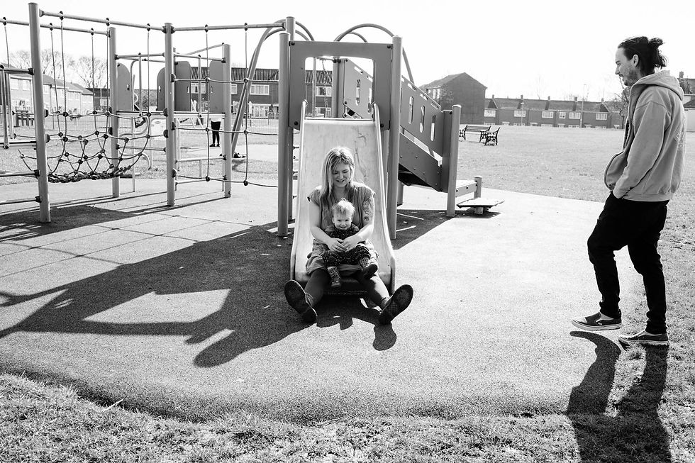 Woman and child sliding down in a playground, smiling. Man watches nearby. Background includes climbing frame and sunny outdoor setting. Family documentary photography Durham and North East. Angela Fenwick Photography
