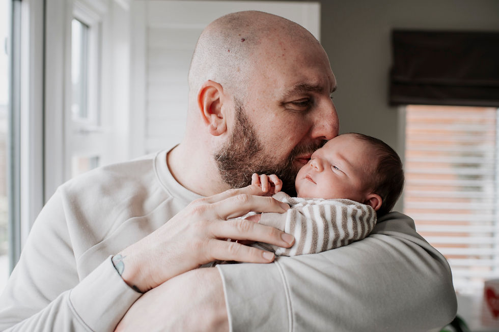 Bald man in a gray shirt gently holds and kisses a newborn wearing a striped babygrow, by a window.