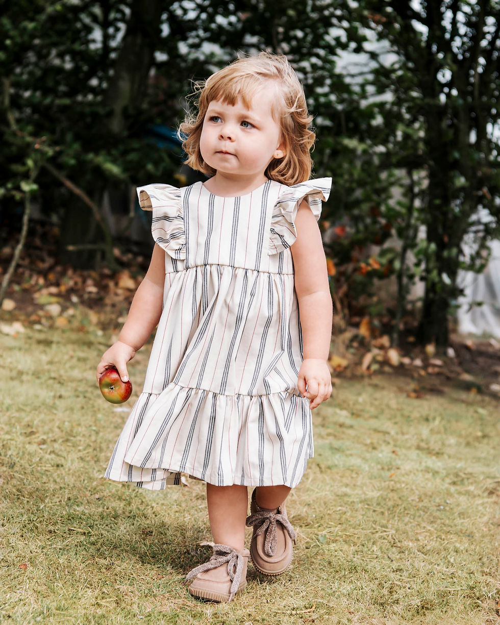 A toddler in a striped dress walks on grass holding an apple, with trees in the background. She appears thoughtful, on a photoshoot in Durham