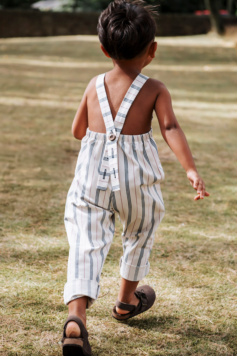 A young child in striped overalls walks on a grassy field, back facing the camera. Sunlight highlights the carefree outdoor setting on an outdoor photo shoot in Duham