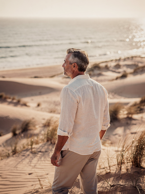 Mann mit grauen Haaren und Bart, steht in den Dünen und blickt auf das Meer. Er trägt ein leichtes, weisses Hemd. Die Dünen sind sandfarben und das Meer ist zu sehen - Mentale Klarheit finden – Ruhe im Kopf in der Natur