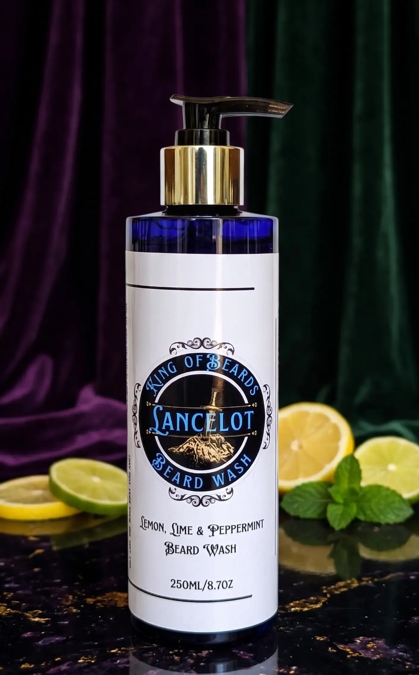 A 250ml blue glass bottle of Lancelot Beard Wash with a gold pump, styled on a dark marble surface with fresh lemon and lime