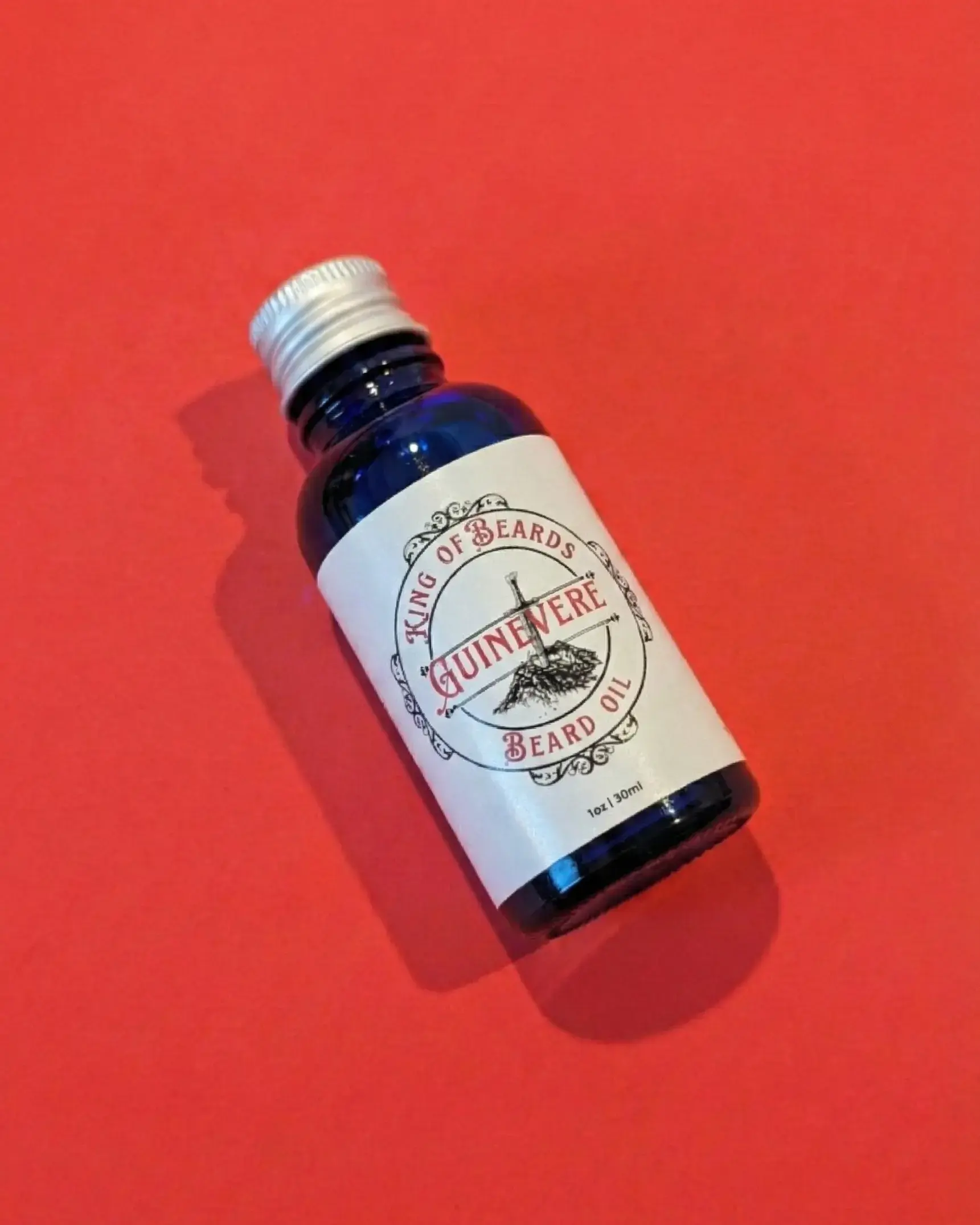 An angled shot of a blue glass King of Beards Guinevere Beard Oil bottle with a silver cap on a rich red background