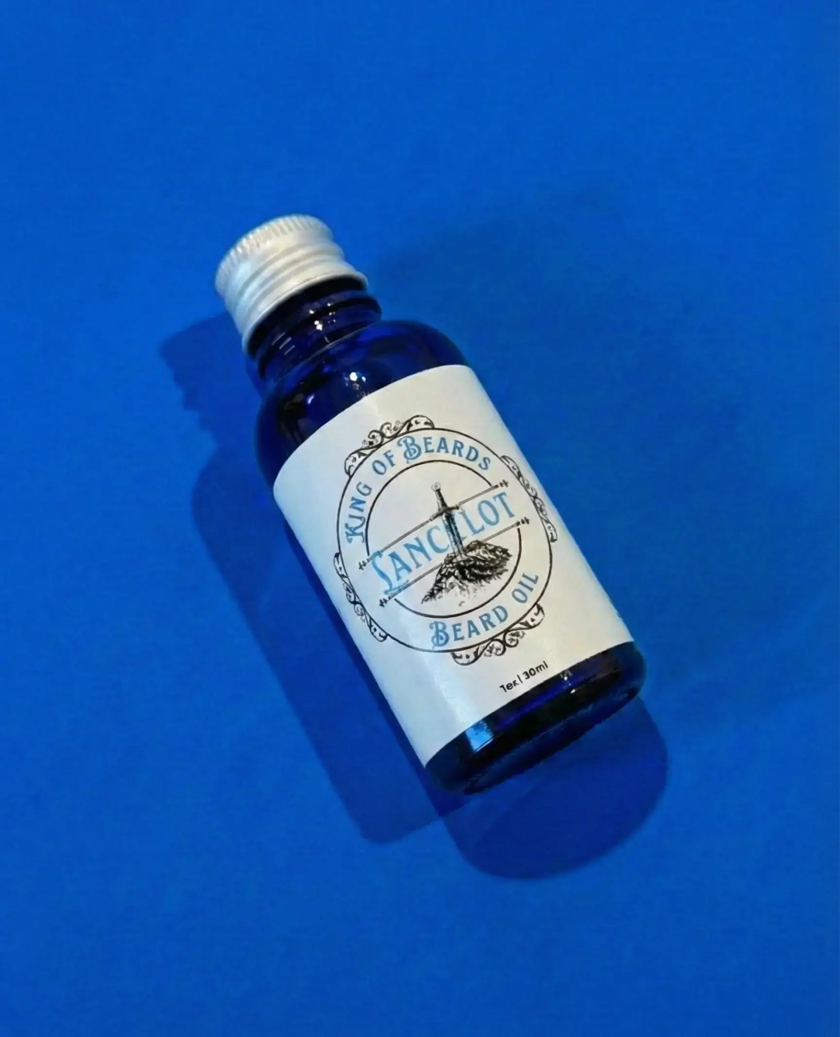 n angled shot of a blue glass King of Beards Lancelot Beard Oil bottle with a silver cap on a vibrant blue background.