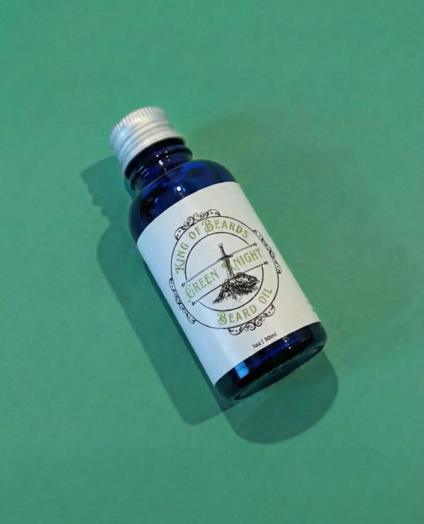 An angled shot of a blue glass King of Beards Green Knight Beard Oil bottle with a silver cap on a green background.