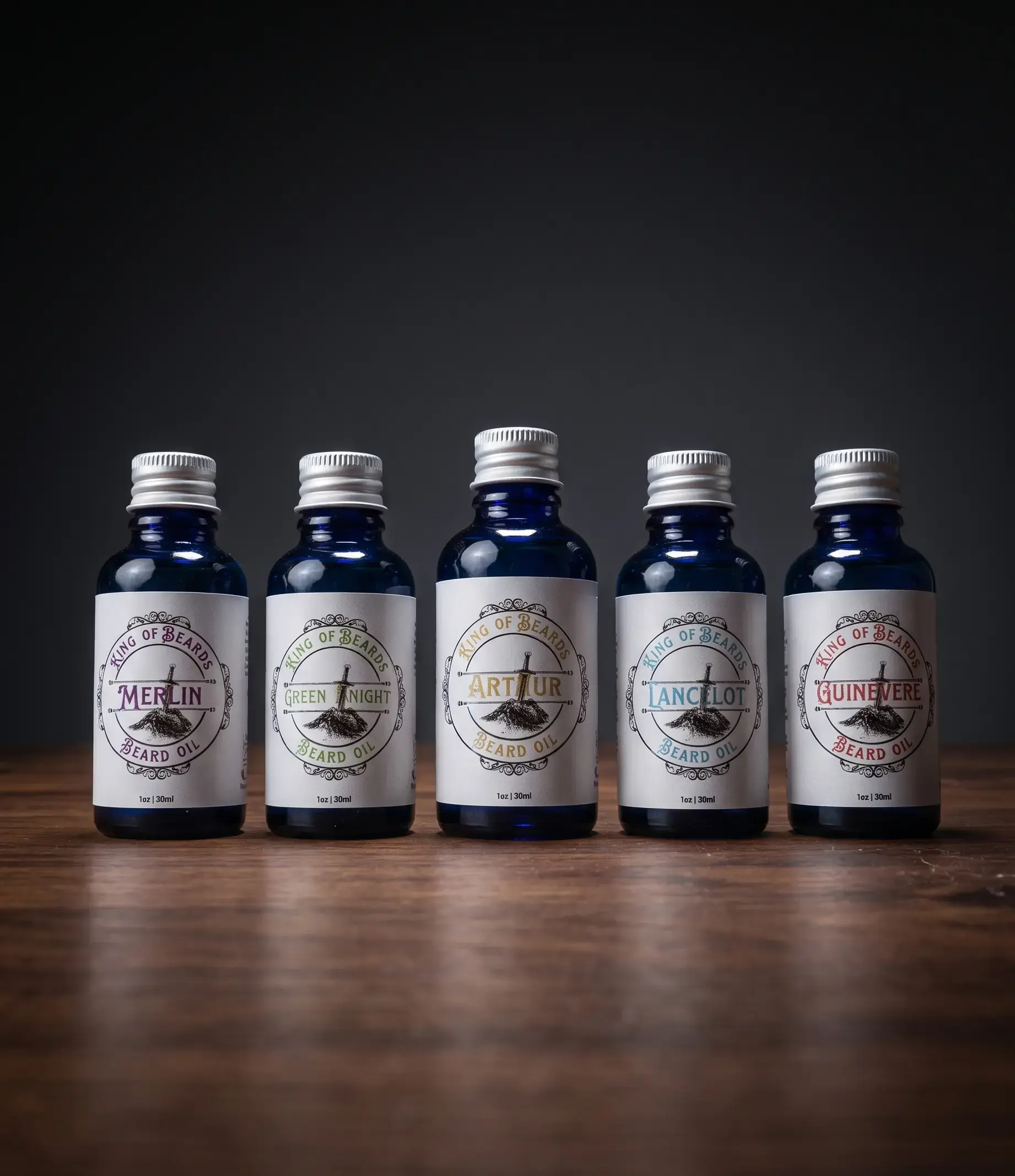 Five blue glass bottles of King of Beards beard oil—Merlin, Green Knight, Arthur, Lancelot, and Guinevere—standing upright