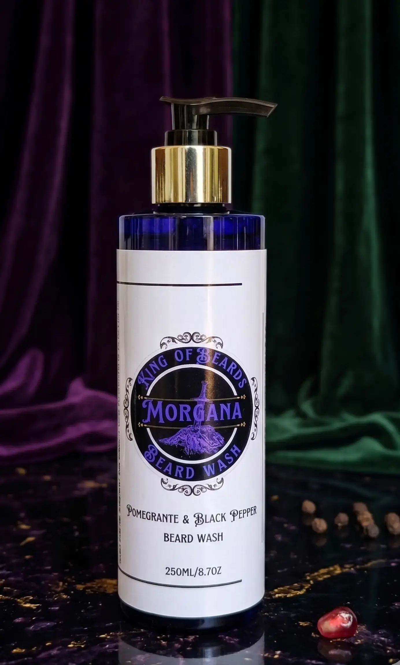 A 250ml bottle of Morgana Beard Wash with a gold pump cap, surrounded by fresh pomegranate seeds and whole black peppercorns