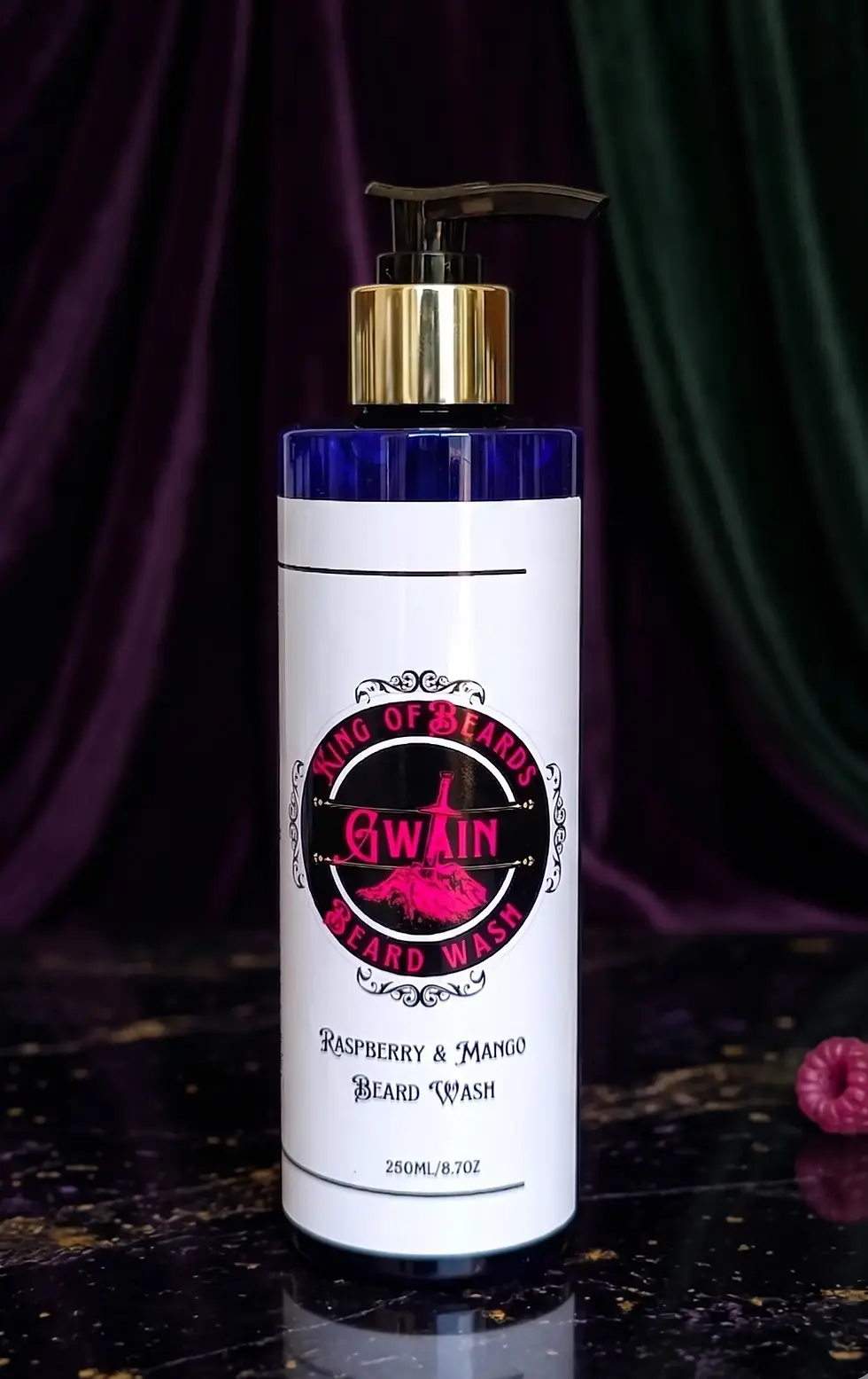 A 250ml blue glass bottle of Gwain Beard Wash with a gold and black pump, set against a dark purple and green velvet curtain 