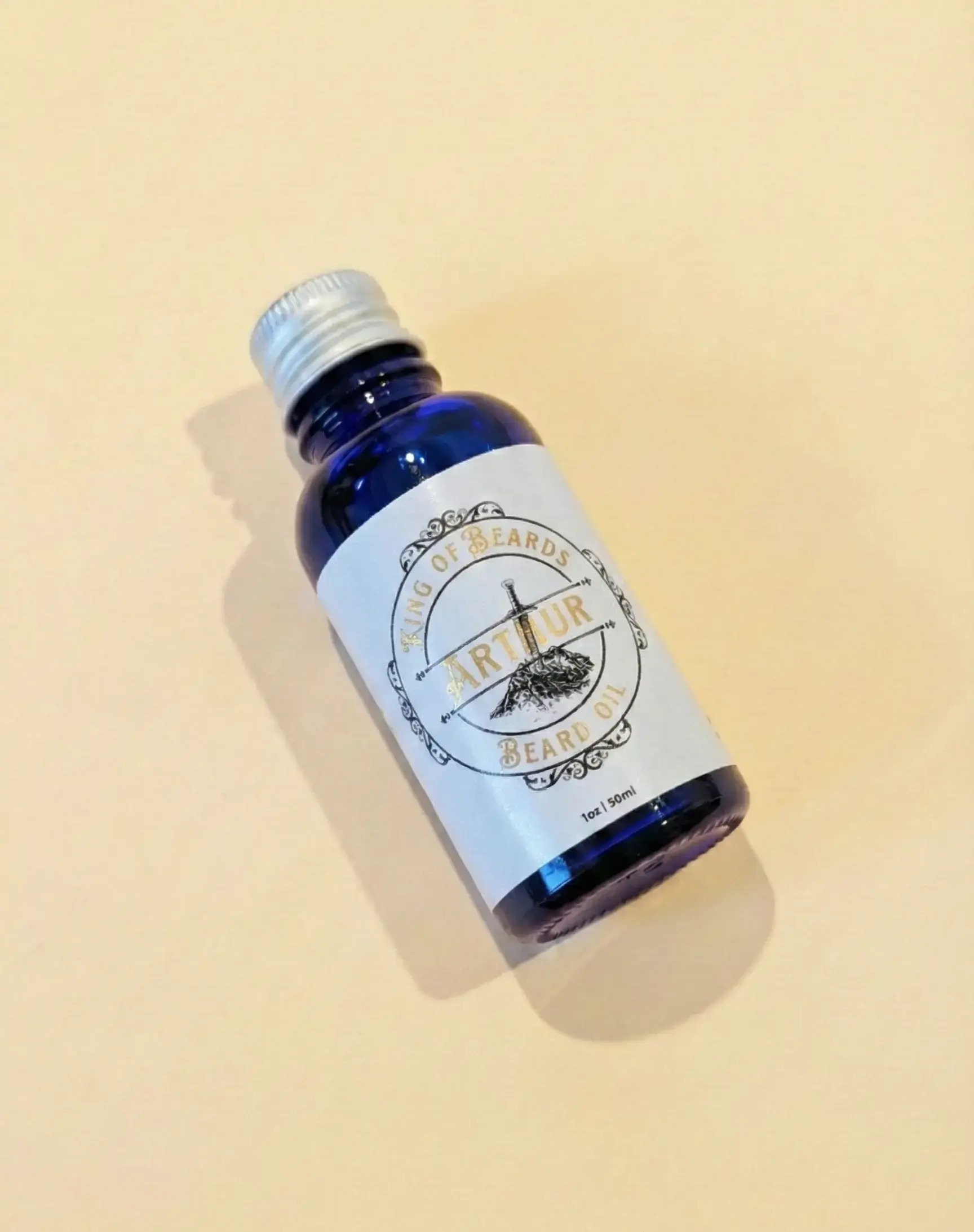An angled shot of a blue glass King of Beards Arthur Beard Oil bottle with a silver cap on a warm cream background
