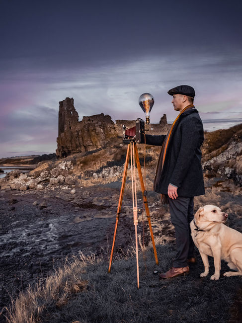 gentleman dresed in vintage attire taking photos with a vintage bellows camera on a woden tripod while his young Labrador sits beside him.