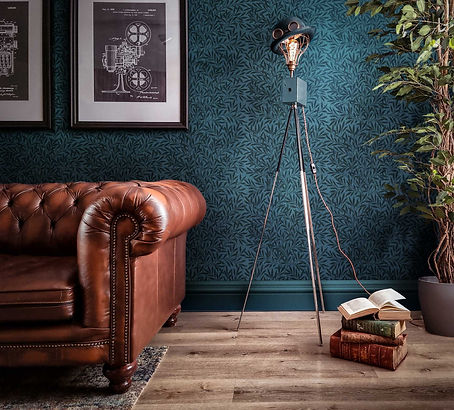 A living room designed by Godwin Vintage with leather Chesterfield sofa, William Morris wallpaper, and an upcycled vintage camera tripod lamp