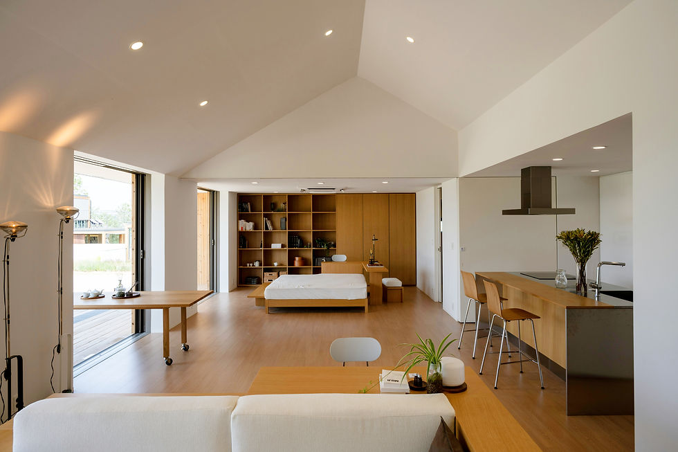 Modern open-plan room with a wooden floor, white walls, bed, bookshelves, and a kitchen island with stools. Natural light streams through open doors.