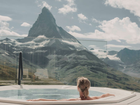 Woman relaxing in a jacuzzi overlooking snowy mountain peaks under a blue sky. Stone wall to the side. Peaceful and scenic setting.