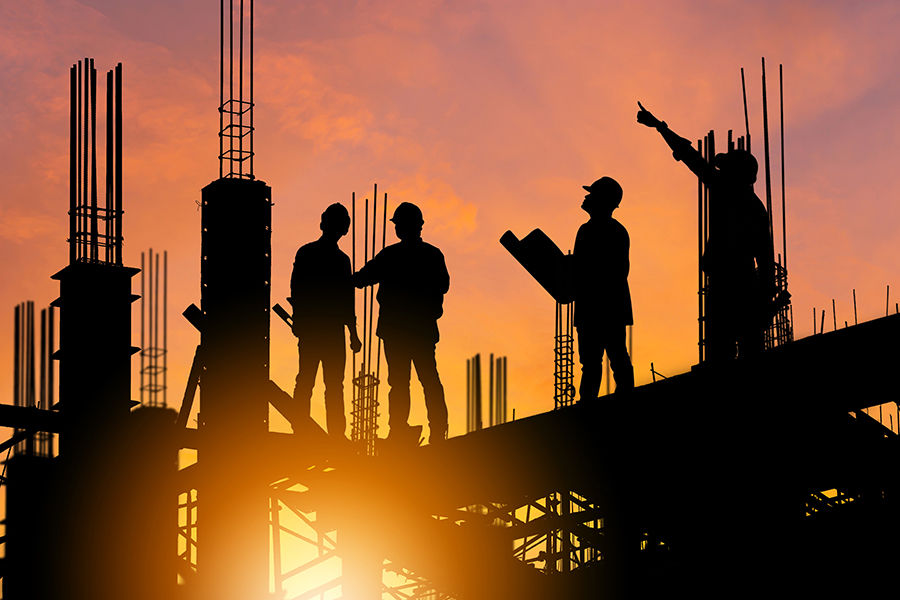 Silhouetted construction workers on site at sunset, holding plans, pointing, and discussing amidst metal beams, with an orange sky backdrop.