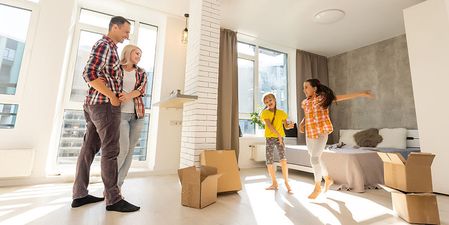 Family in a bright room with moving boxes. Parents smile by windows; kids joyfully dance near a bed. Casual clothes, warm atmosphere.