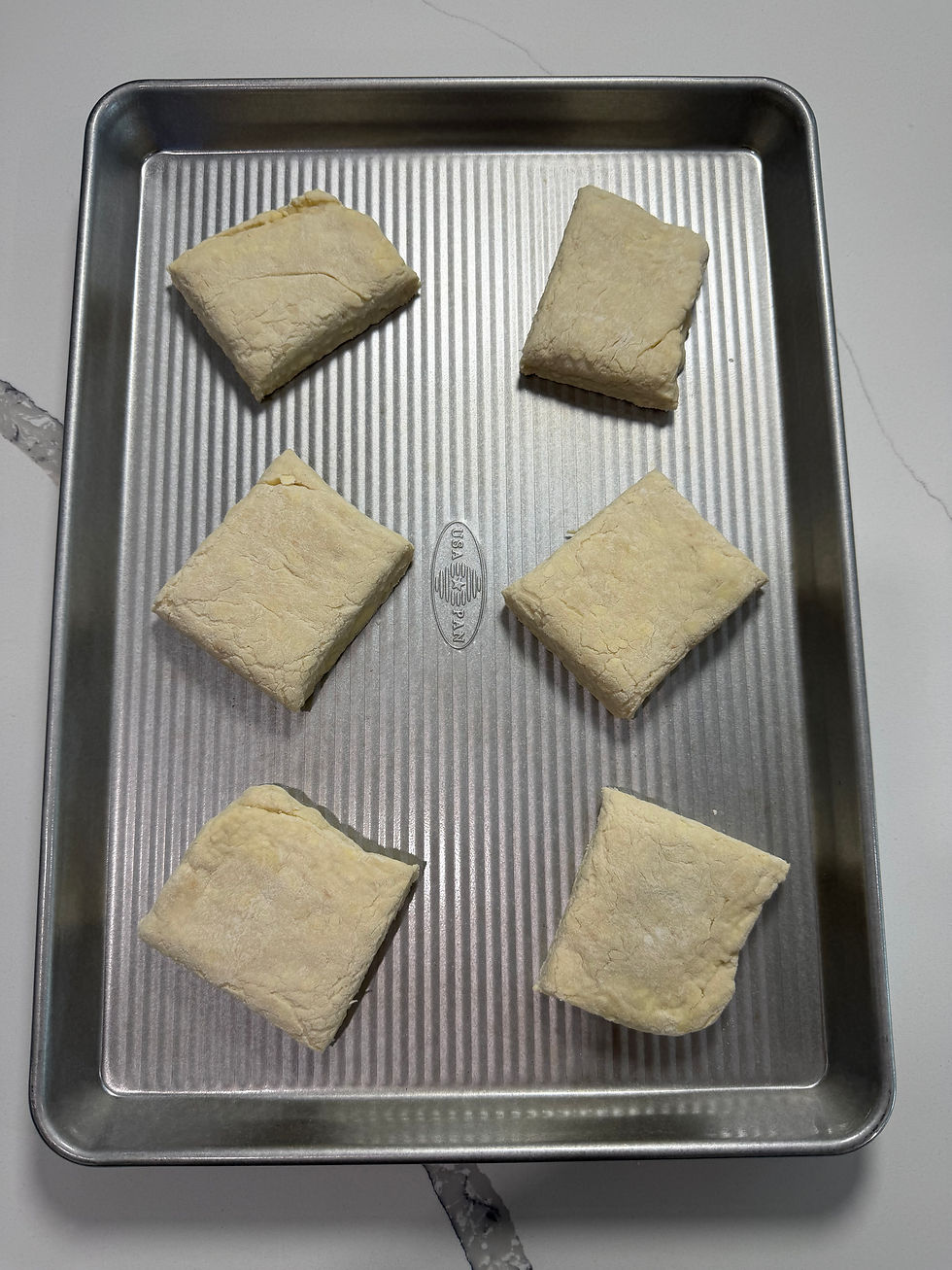 Six square dough pieces on a ribbed USA PAN baking sheet, ready to bake, on a white countertop.