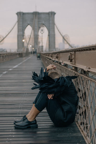 Man sitting on the Brooklyn Bridge boardwalk with black feather wings, moody grey sky and bridge cables above him, NYC editorial portrait