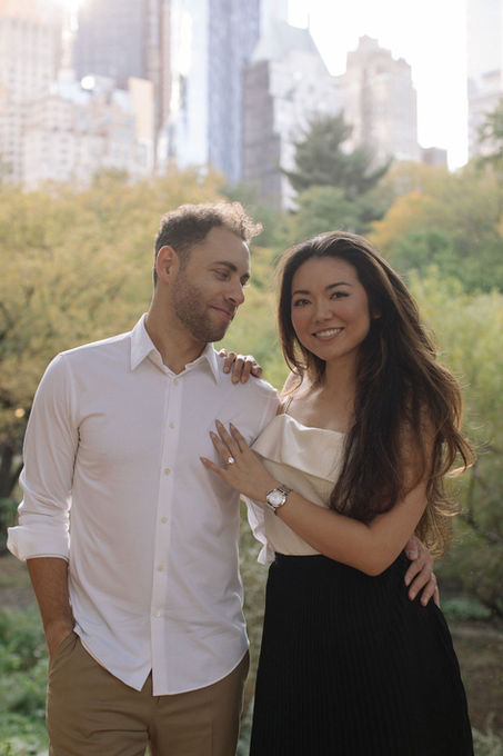 Couple embracing and smiling in Central Park, NYC skyline visible through the autumn trees behind them