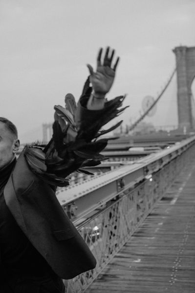 Black-and-white editorial portrait of man with large black feather wings raised on the Brooklyn Bridge boardwalk, NYC conceptual shoot