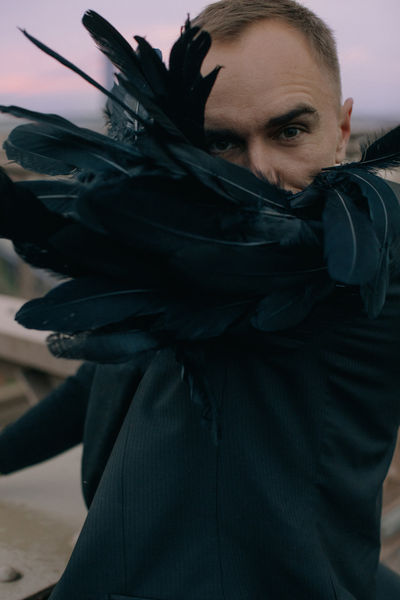 Close-up of man with black feather wings pressed against his face, intense gaze, dark editorial portrait NYC Brooklyn Bridge