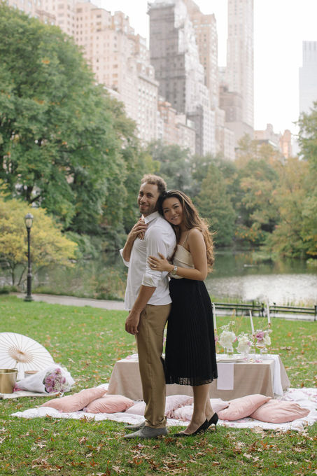 Wide shot of just-engaged couple standing near the Lake in Central Park, NYC skyline in background, golden light