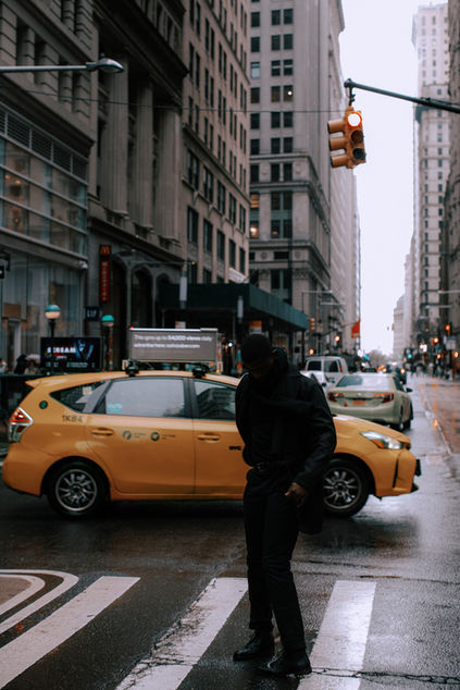 Street portrait of a man in a black coat crossing a rain-soaked Midtown Manhattan intersection with a yellow taxi behind him