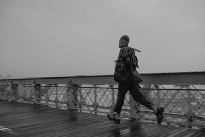 Black-and-white wide shot of man running on the Brooklyn Bridge boardwalk with black feather wings, mist and empty bridge behind him, NYC