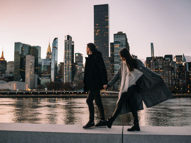 Wide shot of couple on the Roosevelt Island waterfront promenade at dusk, full Midtown Manhattan skyline glowing behind them, NYC