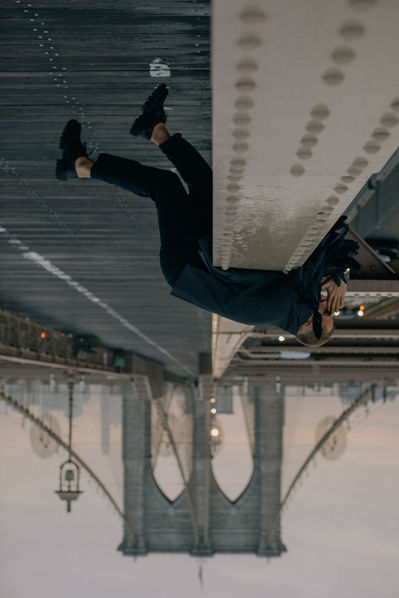 Inverted black-and-white wide shot of man with feather wings upside down on the Brooklyn Bridge, Manhattan Bridge visible in background, NYC