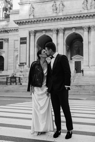 Couple kissing on a Midtown crosswalk, bride in leather jacket over silk gown, New York Public Library behind them