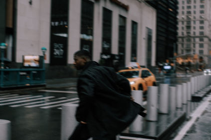 Dynamic motion-blur street portrait of a man in a black coat running past yellow taxis on a rainy Manhattan street