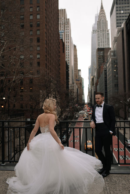 Bride in flowing ball gown and groom in tuxedo posing on a Midtown Manhattan terrace, NYC skyline and skyscrapers at dusk behind them