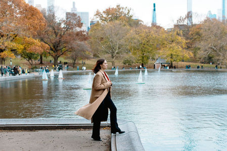 Wide editorial portrait of young woman in camel coat and red top beside the Conservatory Water in Central Park, toy sailboats and autumn trees behind her, NYC