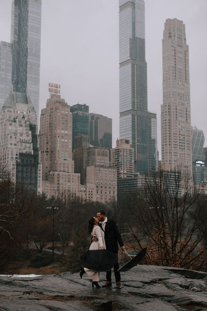 Couple kissing on a rock in Central Park, NYC skyline with luxury towers rising dramatically behind them, winter light