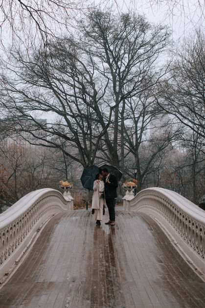Couple walking together across Bow Bridge in Central Park on a rainy day, bare winter trees and soft light around them, NYC engagement