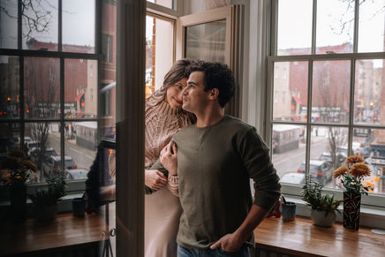 Couple leaning together by a tall apartment window in NYC, Greenwich Village street and buildings visible outside, warm light