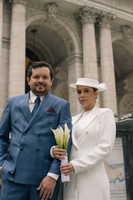 Couple posing beside the stone columns of the New York Public Library, bride in white boater hat holding calla lilies, groom in blue double-breasted suit, NYC