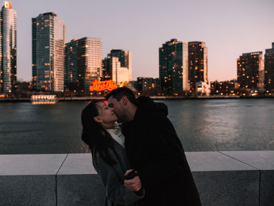 Couple kissing at the Roosevelt Island waterfront at dusk, Midtown Manhattan skyline glowing pink and orange behind them, NYC engagement