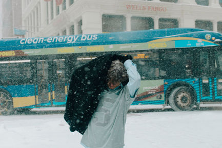 Young man pulling jacket over head in heavy snowfall on a Midtown Manhattan street, NYC bus and Wells Fargo in background, blizzard editorial