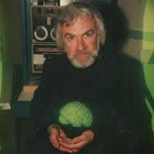 Hombre con barba sostiene cerebro verde brillante. Fondo con equipo técnico. Ambiente enigmático y colores oscuros.