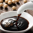 Soy sauce pouring into white bowl with soybeans in background.