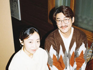 Two people smiling indoors, seated by a wooden window. The man wears glasses and a patterned sweater; the woman is in a light blouse. Cozy mood.