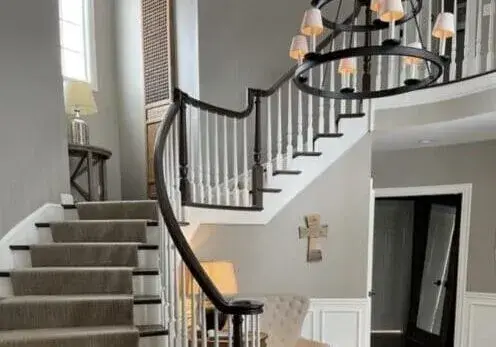 Elegant curved staircase with gray carpet, white banisters, and a chandelier in a bright foyer with a doorway and mirror.