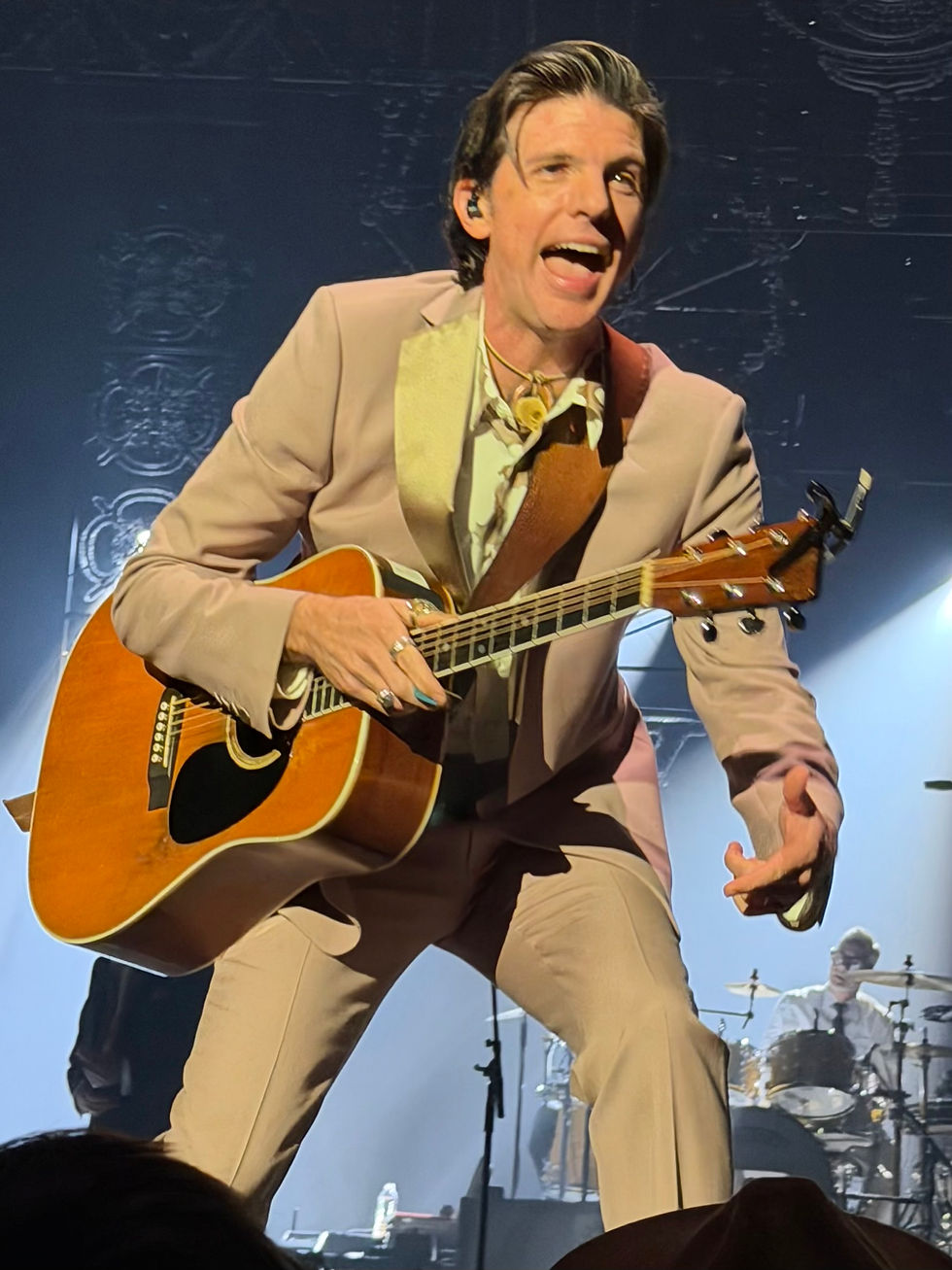 Seth Avett in a tan suit plays acoustic guitar on stage, singing energetically. Drum set in the background. Stage lighting creates an engaging atmosphere.