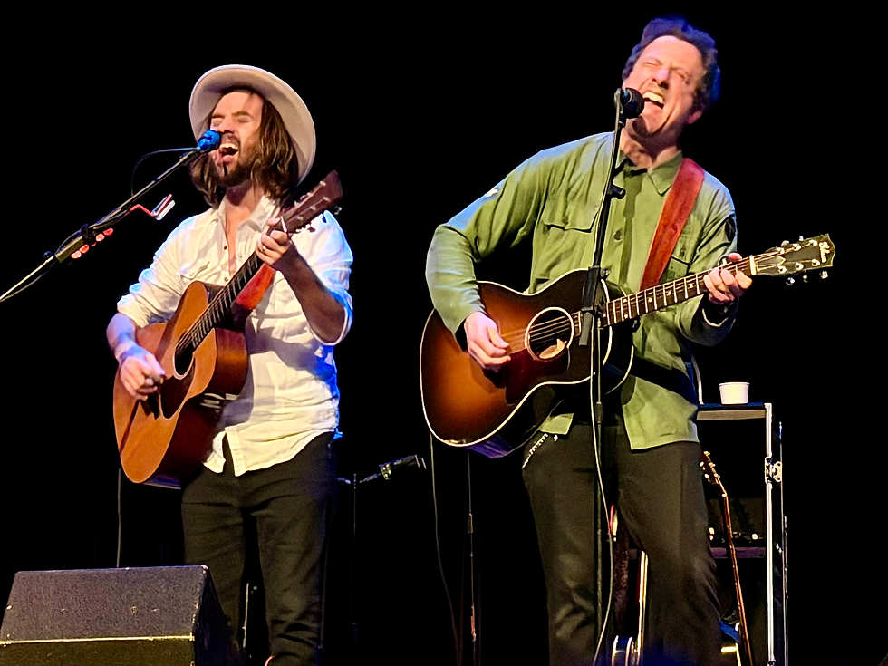 Musicians John Wyman and Will Hoge passionately play guitars and sing on stage. One wears a white shirt and hat, the other in green. Black background.