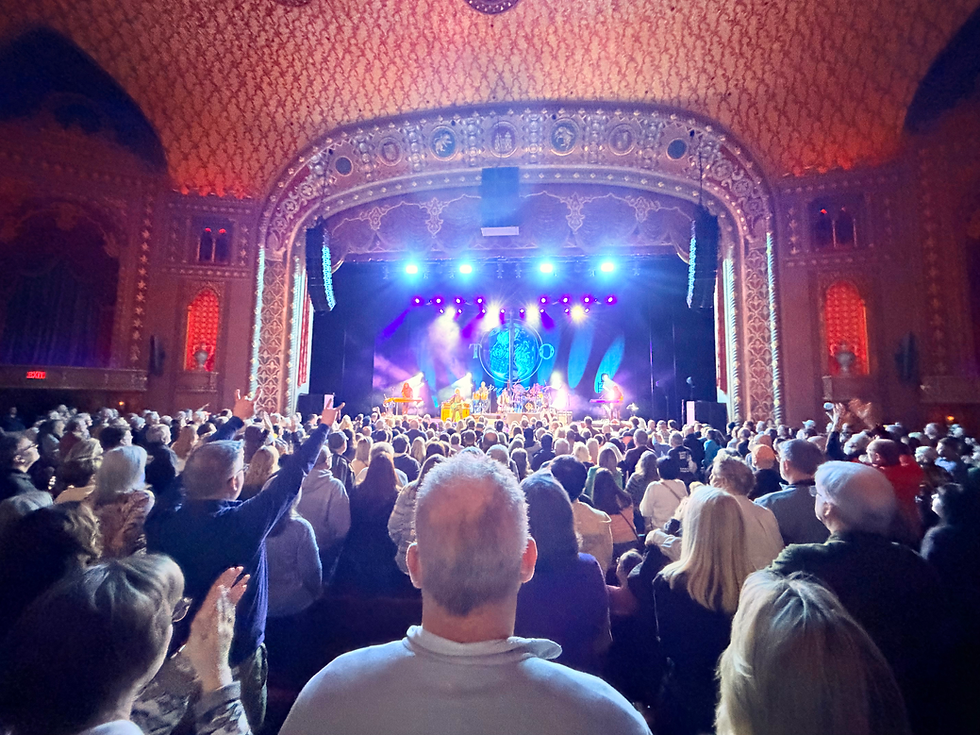 A panoramic view of Toto performing on stage at the Tennessee Theatre, featuring the lights of the theatre around the perimeter of the stage, with the crowd looking on in the foreground.