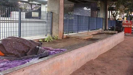 Fotografia urbana mostrando um morador de rua deitado em uma calçada junto a um muro baixo com grades azuis. Ao lado, há uma área verde com vegetação rasteira. Ao fundo, veem-se estruturas de edifícios, contrastando a organização urbana com a realidade da situação de rua