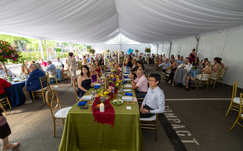 An arrangement of square and round tables placed under a white tent with flowers, linens, table settings, and people sitting at the tables.